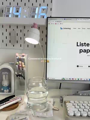 A well-lit desk setup with a digital clock, a computer monitor displaying a website, a desk lamp, a water glass, pens, and a Miffy figurine.