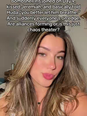 A woman with long, wavy blonde hair is in a selfie-style video. She has a slight smile, and her tongue is sticking out slightly. The background is slightly blurred, suggesting a domestic setting.