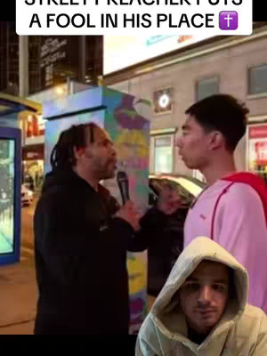 The image shows a street scene with a religious theme. A Black man with dreadlocks is holding a microphone and speaking to another young man. The young man is facing the preacher. In the bottom corner of the scene is another young man, who is also looking at the scene. The text overlay at the top of the image is set over a white background, and uses the words "STREET PREACHER PUTS A FOOL IN HIS PLACE" and has a religious symbol above. There are buildings and street infrastructure visible in the background, with the street scene giving way to a blurry background. The overall lighting appears to be evening or night.