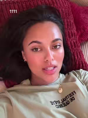 A young woman with dark hair and fair skin is lying down, looking towards the camera. She's on a bed, with a reddish-pink fluffy pillow visible behind her. She wears a light green t-shirt with text in reverse, a necklace, and small hoop earrings. The lighting is soft, and the overall composition is a close-up, focused on her upper body and face.