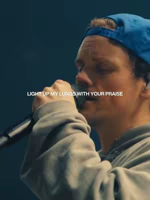 The image captures a man in a low-angle shot, likely during a performance. He is holding a microphone close to his mouth. He has a blue cap, and he is wearing a gray hoodie. The background is a blurred dark teal color.