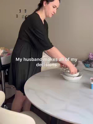 A woman wearing a black dress is standing near a white marble-topped table. She is holding several white bowls, with her hands actively arranging them. The background includes a light-colored wall, and some items on the table adding a touch of domestic scene. The composition is simple with a focus on the woman and her activity.