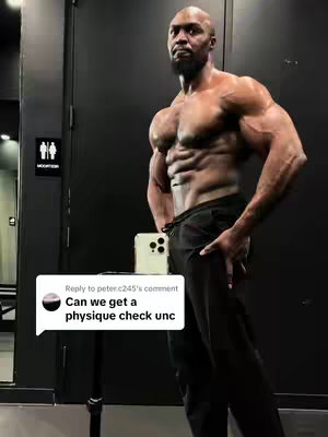 A muscular man poses in a gym, shirtless, with a phone in front of him, holding it as if to take a selfie. The setting is a gym with dark walls and a restroom sign visible in the background.