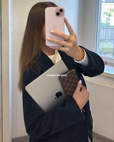 A selfie-style image featuring a person holding a pink phone in front of their face, obscuring it. They also hold a silver laptop and a brown wallet, suggesting a student lifestyle, complemented by the text overlayed on the bottom, which says student life lately. The background is a bright indoor setting.