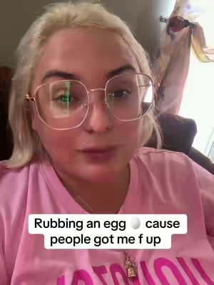 A medium shot features a person with fair skin, blonde hair, and large-framed eyeglasses. They are wearing a pink t-shirt and a delicate pendant. The person is looking directly at the camera with a slight pout. Text overlayed at the bottom states, "Rubbing an egg 🥚 cause people got me f up."