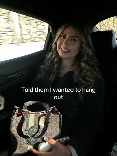 A woman with long, wavy hair smiles while sitting in a car. She looks directly at the camera with a friendly expression. The interior of the car is visible, with a leather-like handbag.