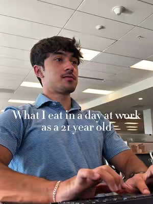 A young man wearing a blue collared shirt is seated at a desk, typing on a black keyboard. The background includes a white ceiling with recessed lighting. Text overlayed on the image reads, "What I eat in a day at work as a 21 year old".