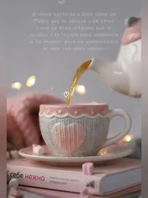 A close-up shot presents a cup with a heart design, being filled with a beverage from a teapot. The scene is surrounded with soft, pink and white tones, with the cup resting on a stack of books.