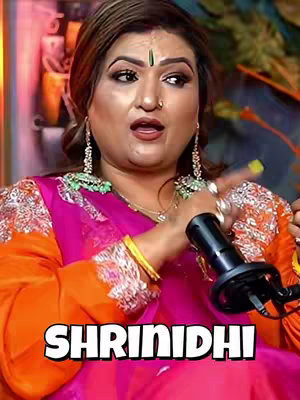 A woman is sitting in front of what appears to be a blurred background. She's looking towards the left side of the frame, with a microphone in her right hand. She's wearing a pink and orange outfit with ornate earrings and a bindi on her forehead. The words "SHRINIDHI" are displayed in bold, white text at the bottom of the image.