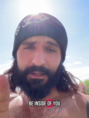 A man with long hair and a beard is shown in a selfie-style shot. He is wearing a black beanie with a logo and his upper body is visible. The background is a bright blue sky.