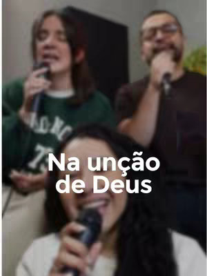 Image shows three people with microphones, singing or speaking, with motion blur. The focus is on the bottom person who is smiling. Text overlayed on the image reads 'Na unção de Deus'.