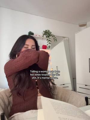 A woman with long dark hair, wearing a brown sweater, is sitting on a bed with her arms covering her face. The background shows a white room with a mirror, shelves with plants, a white cabinet, and a book is visible in the foreground.