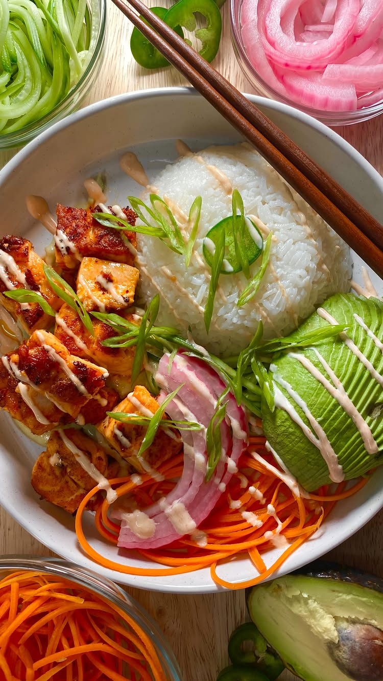 A top-down shot of a gourmet salmon bowl, featuring grilled salmon, white rice, avocado, carrots, pickled red onions, and a drizzle of sauce, all artfully arranged in a white bowl resting on a wooden surface. Chopsticks are casually laid across the bowl, and supplementary ingredients like sliced cucumbers and jalapeños are in the background, with the overall composition appearing appetizing and photogenic.