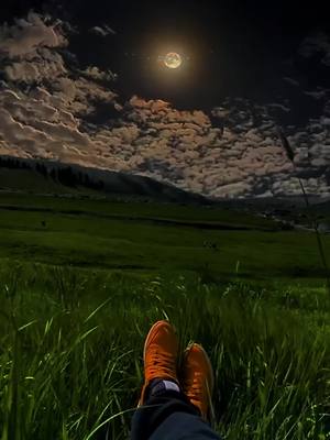 A night scene with low angle view of a person wearing orange shoes sitting in grass, with a full moon up above in the cloudy night sky.