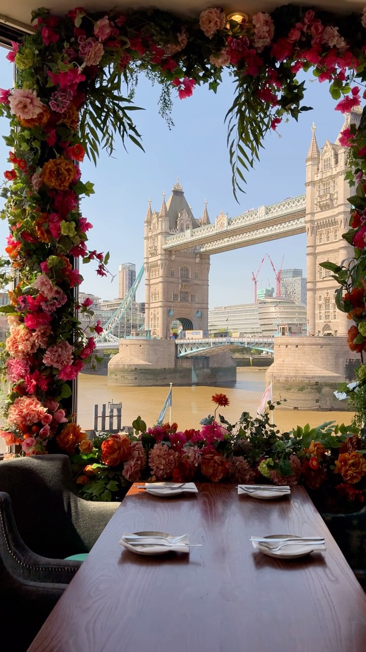A photograph of a restaurant window with a view of the Tower Bridge in London. The window frame is filled with an abundance of colorful flowers, framing the bridge. The bridge is visible in the background with the river and other buildings. A wooden table is in the foreground with plates and silverware.