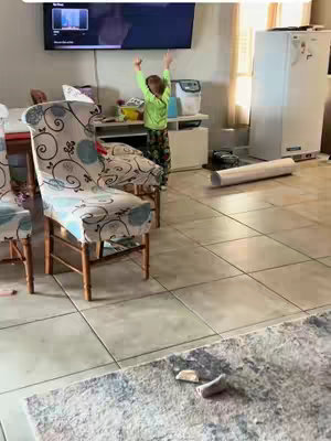 A young child stands in a living room with arms raised in front of a TV. The room has tile flooring, chairs with patterned covers, and a rug with scattered objects.