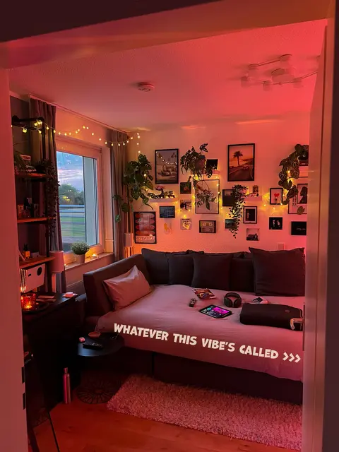 A cozy bedroom interior is captured through a doorway. Warm lighting illuminates the room, highlighting the bed, which is the central element, made up with pillows. Wall art, plants, and string lights add to the ambiance of the room. On the bed a phone, a controller and a headphone are visible.