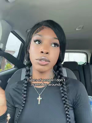 A woman with dark braids, wearing a black long-sleeved shirt and a cross necklace, poses while looking into the distance. Text overlay at the bottom. The setting seems to be inside a vehicle.
