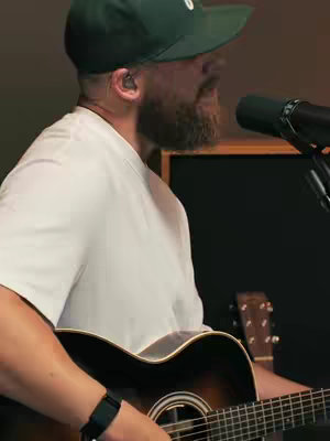 The image features a man in profile, playing an acoustic guitar. He wears a white t-shirt, a green baseball cap, and a black wristwatch. A microphone is positioned in front of him, with a dark background and a second guitar visible in the blurred background. The lighting suggests a studio or indoor setting.
