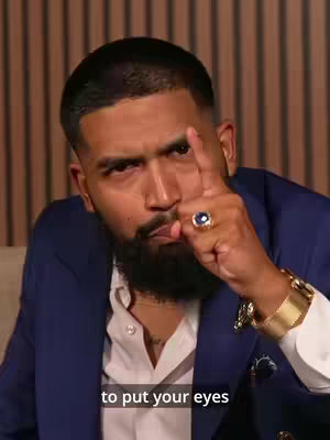 A man with short black hair, a beard, and a navy blue suit looks directly at the camera, pointing one finger upward. He wears a large blue ring and a gold watch. The background is a light brown, vertical-paneled wall.
