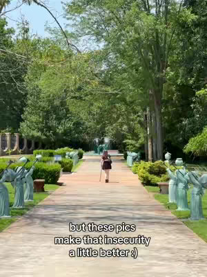 A person with a walking stick is seen from behind, walking on a paved path between manicured hedges and flower statues. The background includes trees and a partly overcast sky.