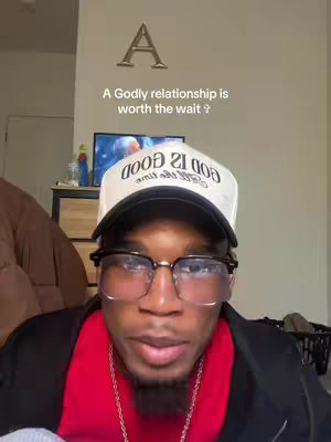 A selfie-style shot of a man with dark skin, wearing glasses, a hat, and a red shirt. He is positioned from the chest up, looking towards the camera with a contemplative expression. The background is slightly blurred, but hints at a light-colored wall and some furniture. The lighting appears to be natural, and the overall composition is straightforward, focusing on the subject.