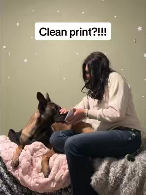 A woman seated on a bed with a dog, holding a small object near the dog's face. The dog appears to be interacting with the object. A text overlay reads, "Clean print??!!" The composition is candid, with a casual, intimate setting.