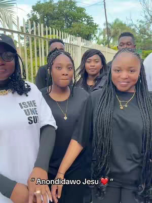 A group of six people, primarily women, are standing outside. The main focus is on the group of young women in the center, who all dressed in similar outfits, while the background features a fence and foliage.