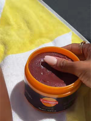 A close-up shot shows a hand with a ring holding an open orange jar of Carroten tanning gel. The jar is resting on a yellow and white striped towel. The gel inside is a dark maroon color, with a finger touching its surface.