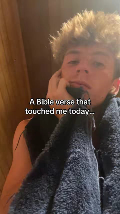 A young man with light brown, messy hair is shown from the chest up, with a dark background. The man is holding a dark blue towel or cloth to his face. White text overlayed in the face states, “A Bible verse that touched me today…”.