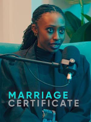 A woman is seated on a teal couch in a studio setting. She has dark hair styled in locs, and is wearing a dark blazer over a dark turtleneck. A microphone is positioned in front of her. Behind her are some plants, a teal wall, and soft studio lighting.