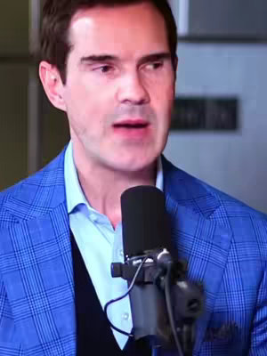 The image is a medium shot of a man speaking into a microphone. He is wearing a blue plaid blazer over a light blue collared shirt and a dark sweater or undershirt. The microphone is a large, professional model placed close to his mouth. The background is slightly blurred, suggesting an interior setting. The lighting appears to be focused on the man. The composition is a standard podcasting setup, with the man positioned centrally framed.