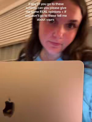 A young woman is looking directly into the camera, with a laptop computer in the foreground. The background has horizontal blinds which are slightly out of focus.