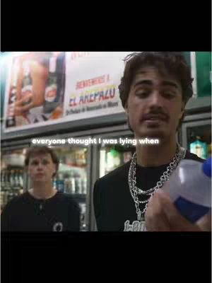 The cover image captures a scene inside what appears to be a convenience store, with two young men in focus. The primary figure, positioned in the foreground on the right, is a man with a mustache and dark hair. He wears a black t-shirt, and multiple silver chains, He's holding a clear plastic bottle, his gaze direct. Behind him, another man with fair skin and short hair is visible. The background features shelves with products, signage with spanish text, and a freezer. The lighting is bright, indicating a well-lit shopping environment.