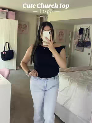 A woman in a black top and light blue jeans is taking a mirror selfie. The image shows her in a room with a bed and other home decor, with text overlayed at the top.