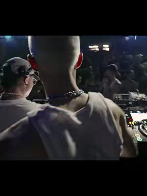 The video cover image shows the back of a person with short, light-colored hair, wearing a white tank top, facing a crowd. Another person wearing a hat is seen from the back on the left. The setting appears to be a concert or a public event with many people in the background, and equipment to the right of the main subject.