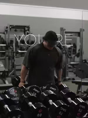 The image shows a man lifting weights in a gym, seen from a slightly low angle. Text overlays the image. The man is wearing a dark outfit and a backward cap. Rows of dumbbells are in the foreground.