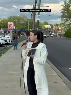 A woman is standing on a sidewalk, outside on a sunny day, holding a microphone and a book. A street is in the background with cars driving.