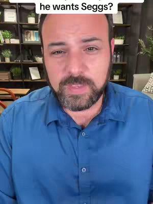 A man with a beard and short dark hair is centered in the frame, wearing a blue button-down shirt. Above his head is a white text overlay. The background is slightly blurred, showing hints of shelves with items and plants.