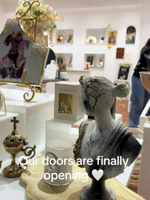 An interior shot showcasing a display of goods with a statue in the foreground. Text overlayed reads 'Our doors are finally opening' with a white heart. The setting includes shelves, boxes a gold tray and ornamental objects. The overall palette is neutral, using soft lighting.