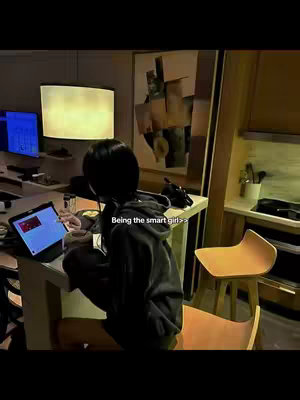 A young woman with dark hair is seen from behind, seated at a desk, engrossed in her laptop. The image is warmly lit with a soft, slightly blurred background featuring elements of a modern home office or study area.
