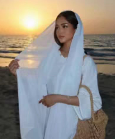 A woman wearing a white head covering and dress stands on a beach at sunset, holding the fabric of her veil. She is facing the camera with a serene expression, and a woven bag hangs from her arm. The warm sunlight casts a golden glow over the scene, with the ocean and sky visible in the background.