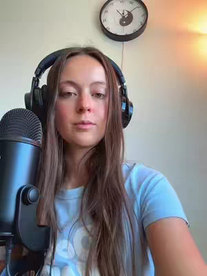A young woman with long brown hair wearing a blue t-shirt and headphones looks directly at the camera while holding a black microphone. A clock with a yin-yang symbol hangs on the wall behind her.