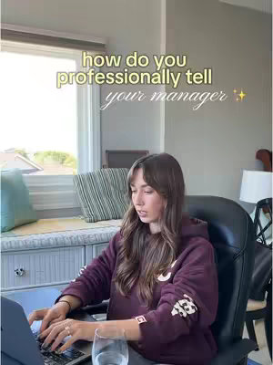A woman sits at a desk indoors, typing on a laptop. The room is naturally lit with a window showing the outside. Overlaid text suggests the video is about asking for advice from a manager.