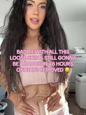 A selfie-style shot of a woman in a pink crop top with dark hair. Text overlays her. The woman's hands are holding up her lower torso, showing the stomach and loose skin. Background shows a white room with some furniture, and a white washing machine.