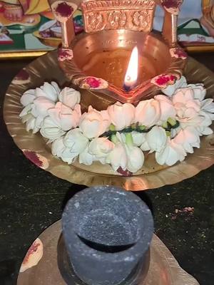 The image features a lit diya lamp placed on a brass plate decorated with white jasmine flowers. Below is a partially visible stone mortar. The lamp, with its flame, is at the center, with the flowers forming a wreath around the rim of the plate.