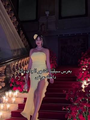 A woman in a yellow, strapless gown is walking down a red-carpeted staircase, framed by ornamental arrangements of red flowers and lit by candles. Text in Arabic is overlaid on the image.