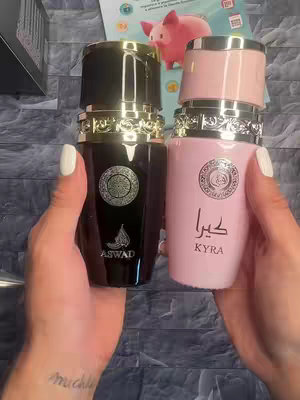 The cover image shows a close-up of a person holding two perfume bottles. The bottles are next to each other in front of light-colored shelves. The person's hands are prominent, with a tattoo visible on one wrist. Both bottles have unique designs. The one on the left is dark with a gold cap and a circular label. The one on the right is pink with a matching pink cap and text in a different language. The background is somewhat blurred.