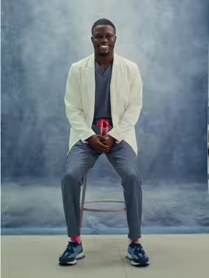 A man in professional attire sits on a stool against a cloudy blue background. He wears a white lab coat over a dark blue V-neck shirt and matching trousers. His hands are clasped in front of him. He has dark skin, is smiling, and wearing blue sneakers and pink socks. The background has a watercolor effect.