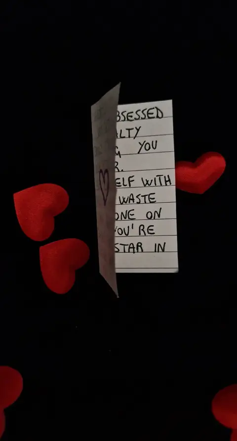 A still life composition with a black background, featuring several plush red hearts scattered across the surface. The main focus is a folded white piece of paper with handwritten text on it. The paper is slightly angled, showing the handwritten message, which is slightly out of focus. The overall layout is simple, with the focus on the message and the hearts.